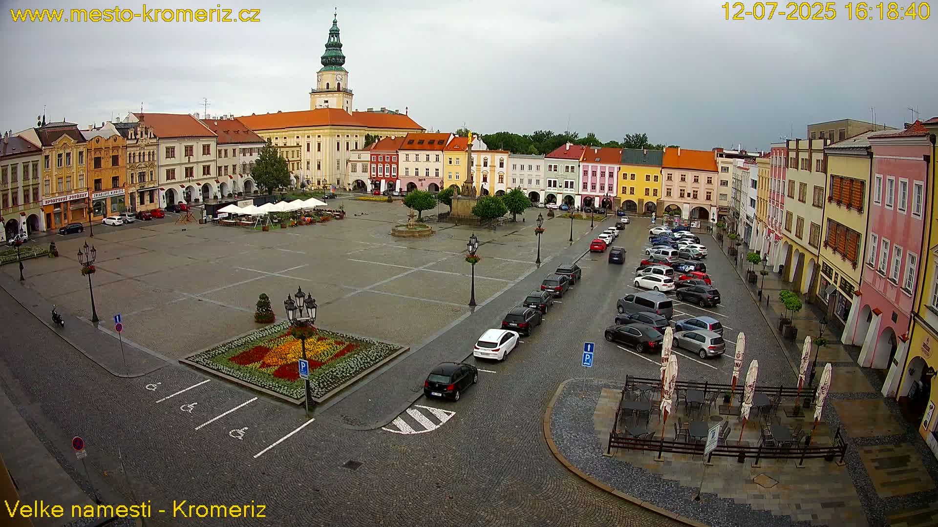A large town square with colorful buildings surrounding it, cars parked along the sides, and a flower garden in the center, under sunny conditions.
