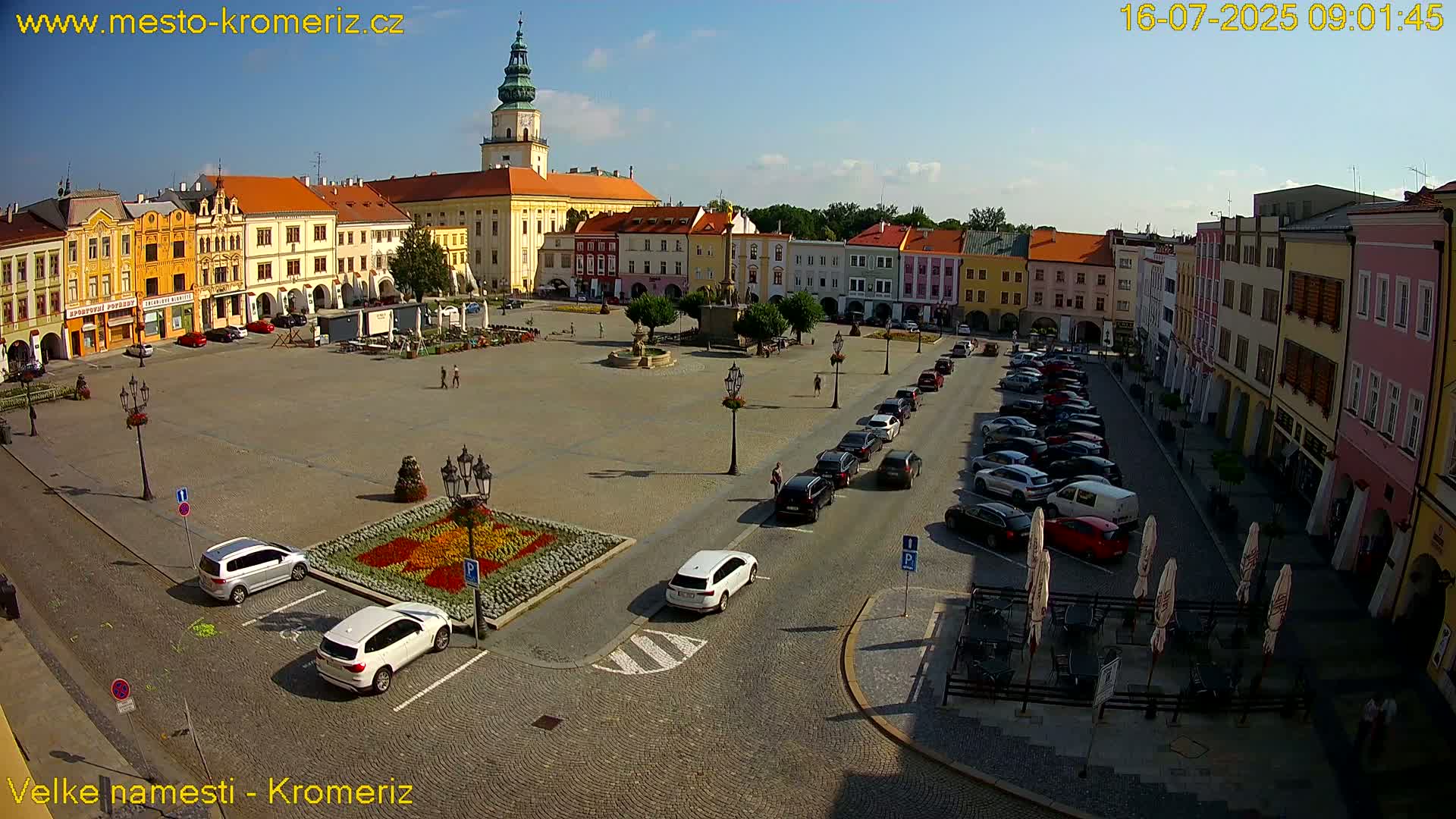 A large town square with colorful buildings surrounding it, cars parked along the sides, and a few people walking around under a clear, sunny sky.