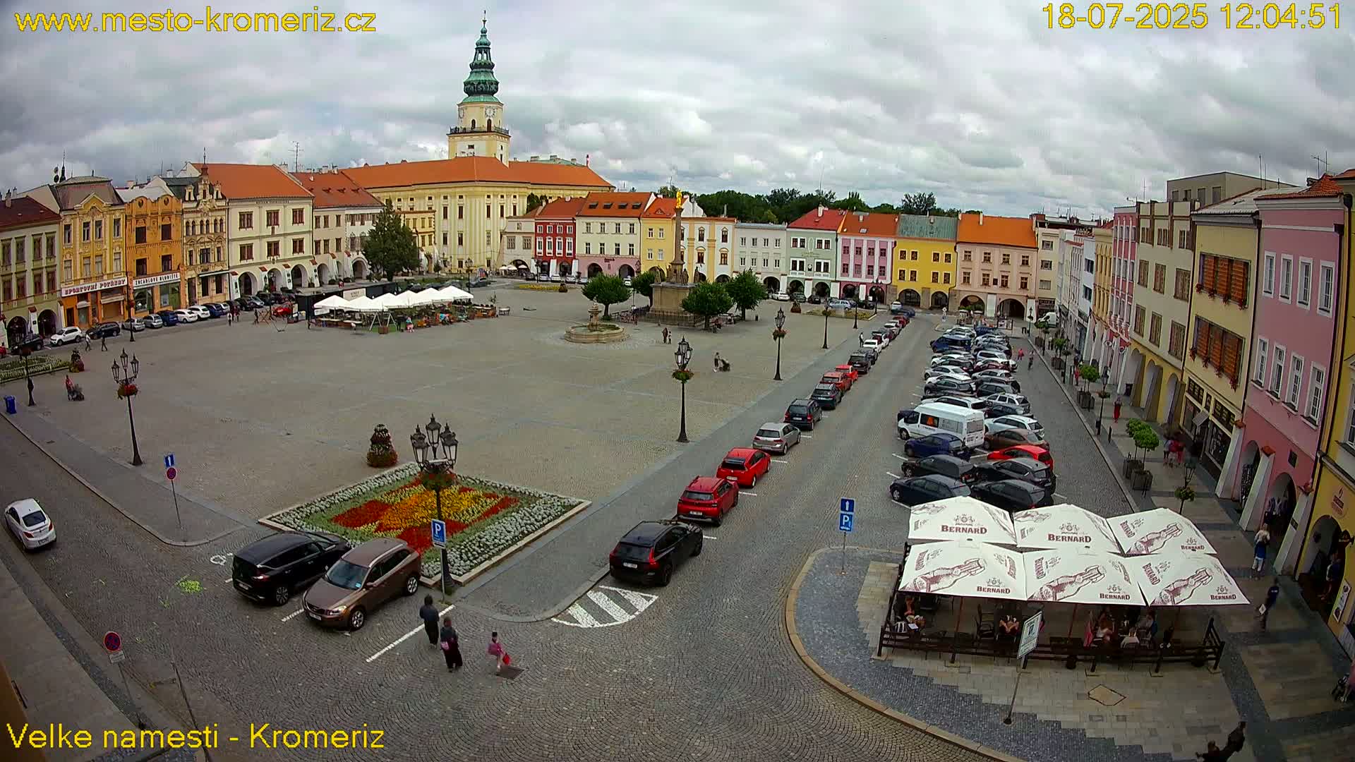 A large town square with colorful buildings surrounding it, under a cloudy sky, features cars parked along one side and a flower garden in the center.