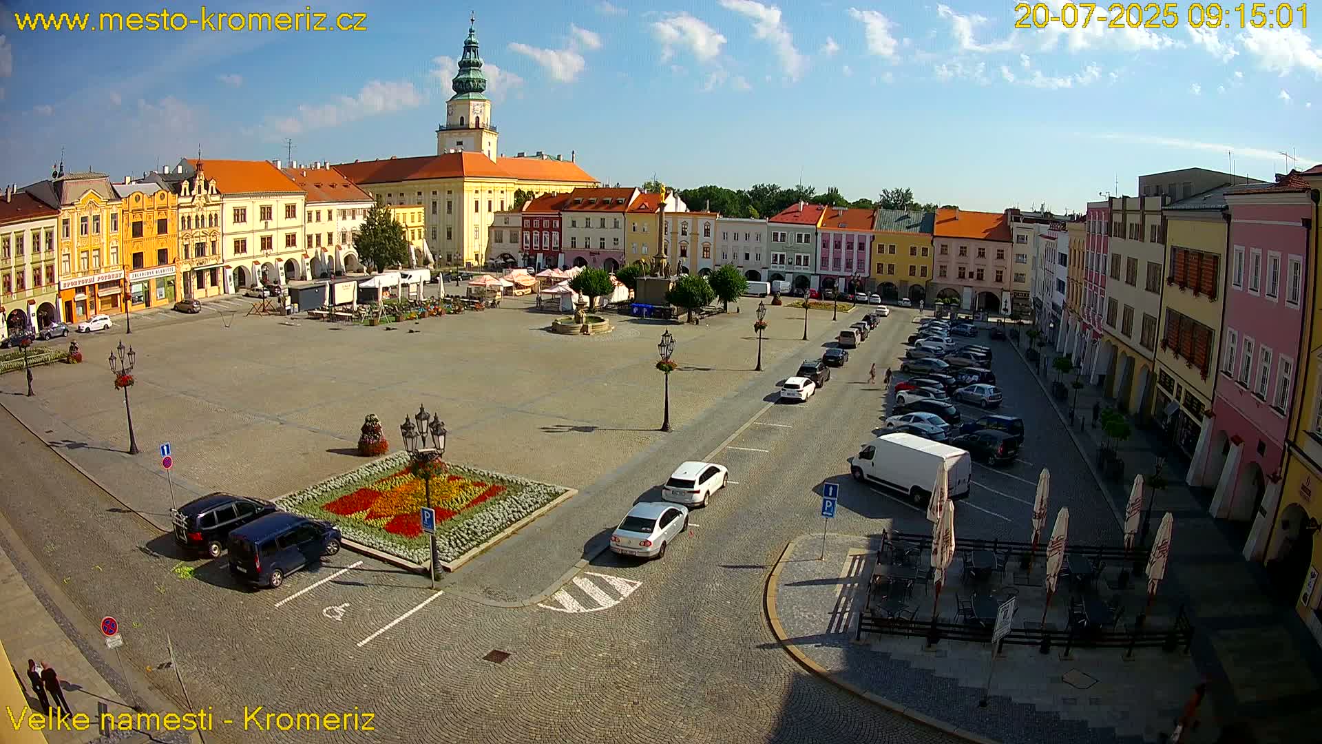A large town square with colorful buildings surrounding it, parked cars, and a flower bed in the center, under a mostly sunny sky.
