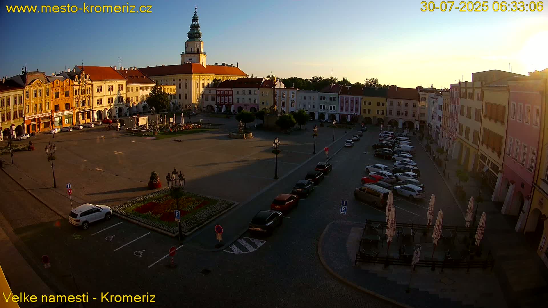 A large town square with colorful buildings surrounding it, parked cars, and a flower bed in the center, under a mostly sunny sky.