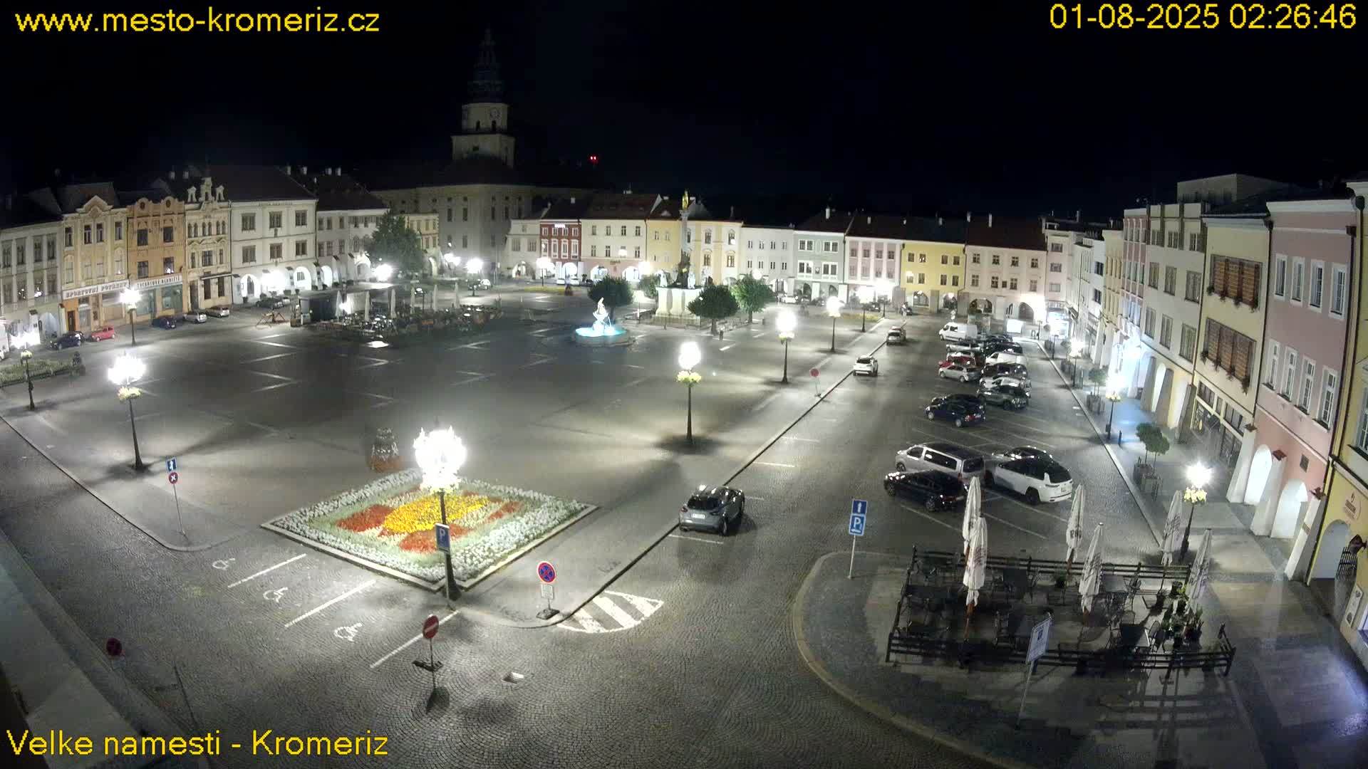 A nighttime, illuminated town square with parked cars, a fountain, and buildings surrounding it.