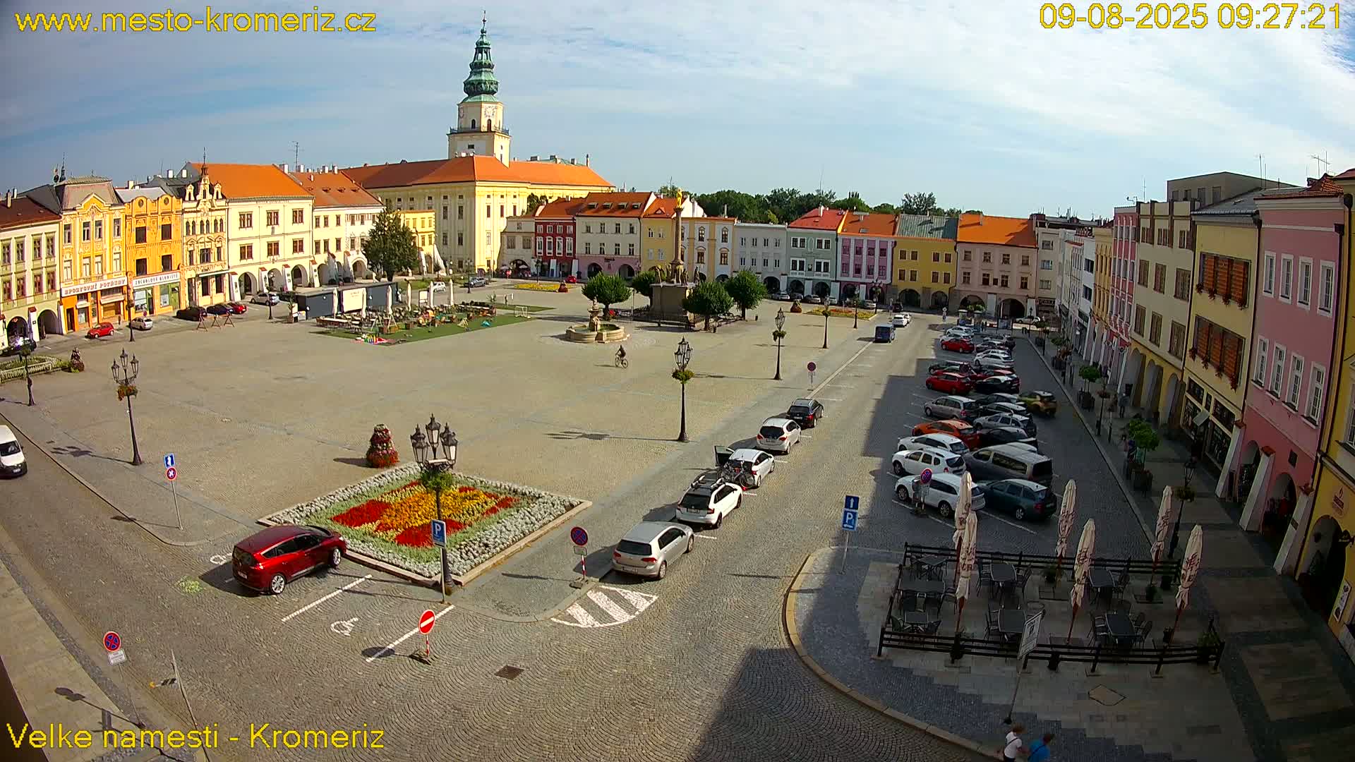 A large town square with colorful buildings surrounding it, cars parked along the edges, and a flower garden in the center, under sunny skies.