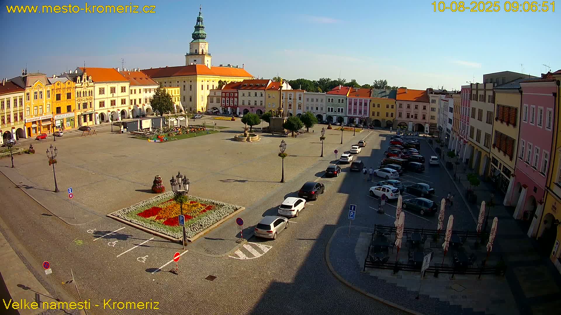 A large town square with colorful buildings, a central fountain, parked cars, and a flower bed, all under a sunny sky.