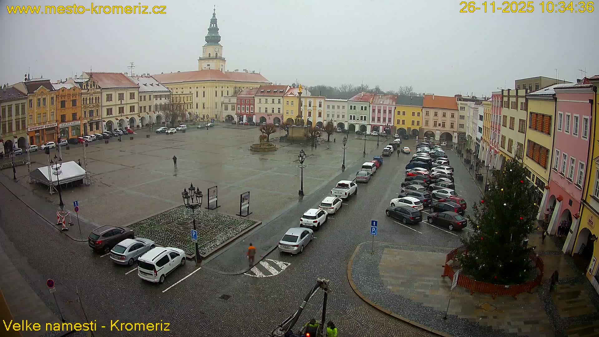 A large, wet cobblestone town square is visible on an overcast and damp day, surrounded by colorful historic buildings, a prominent clock tower, a central fountain, numerous parked cars, a decorated Christmas tree, and a few pedestrians.