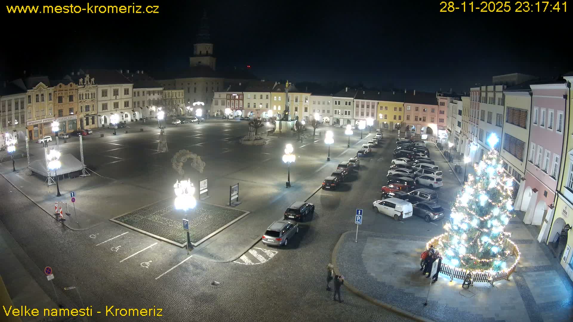 A well-lit European town square at night features a large, glowing Christmas tree, surrounded by historic buildings, numerous parked cars, streetlights, and a few scattered pedestrians, all under clear, dark skies.