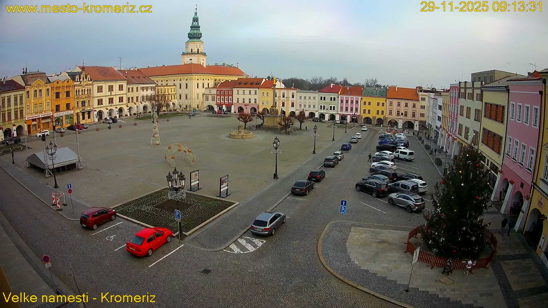 A wide, historic European town square, featuring colorful buildings, a central fountain, scattered Christmas decorations including a large tree and light arches, parked cars, and a prominent green-domed tower in the background, all under an overcast sky.