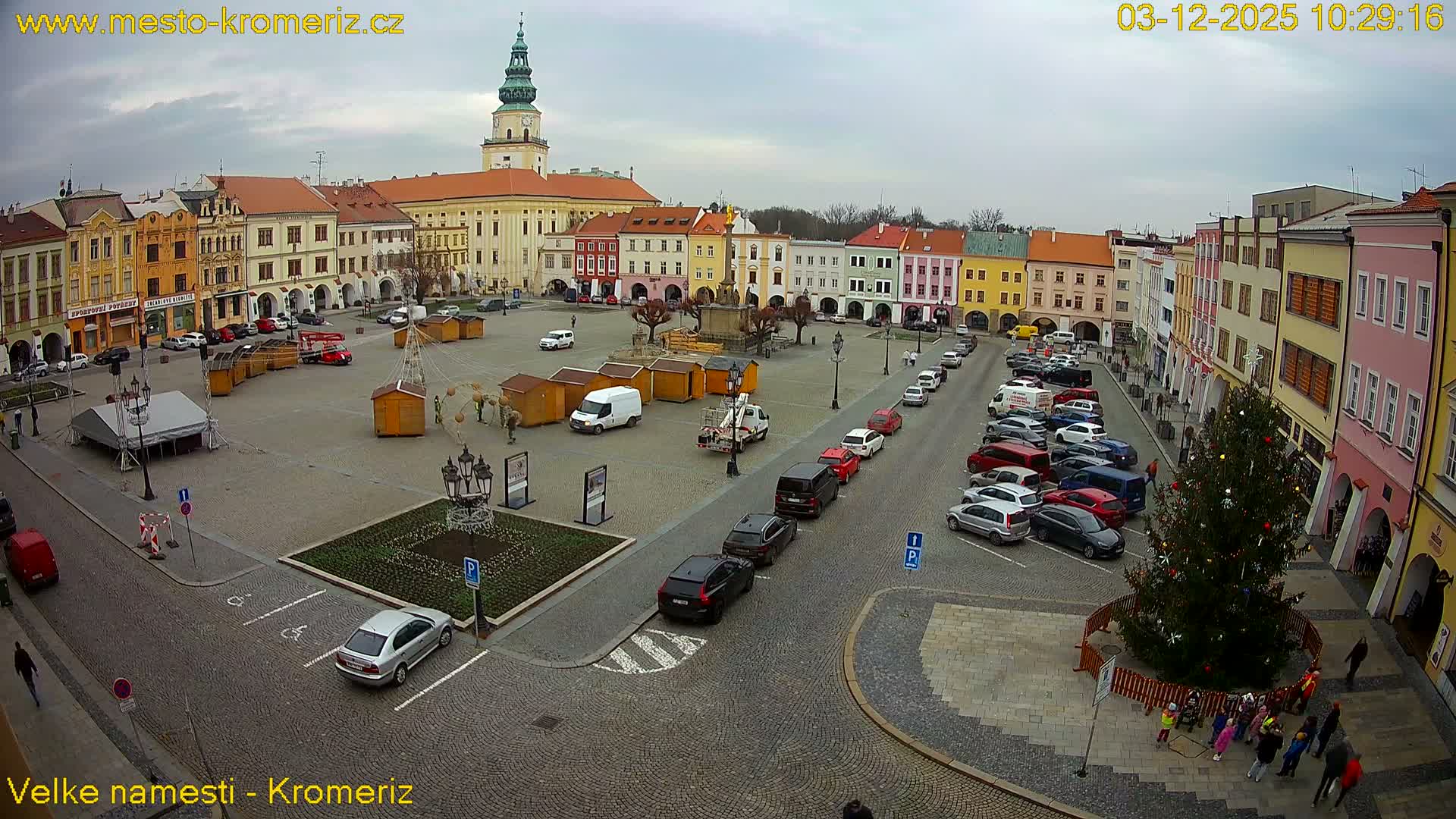 An overcast sky hangs over a European town square lined with colorful historic buildings, featuring a central monument, numerous parked cars, market stalls, and a decorated Christmas tree with people gathered around it.