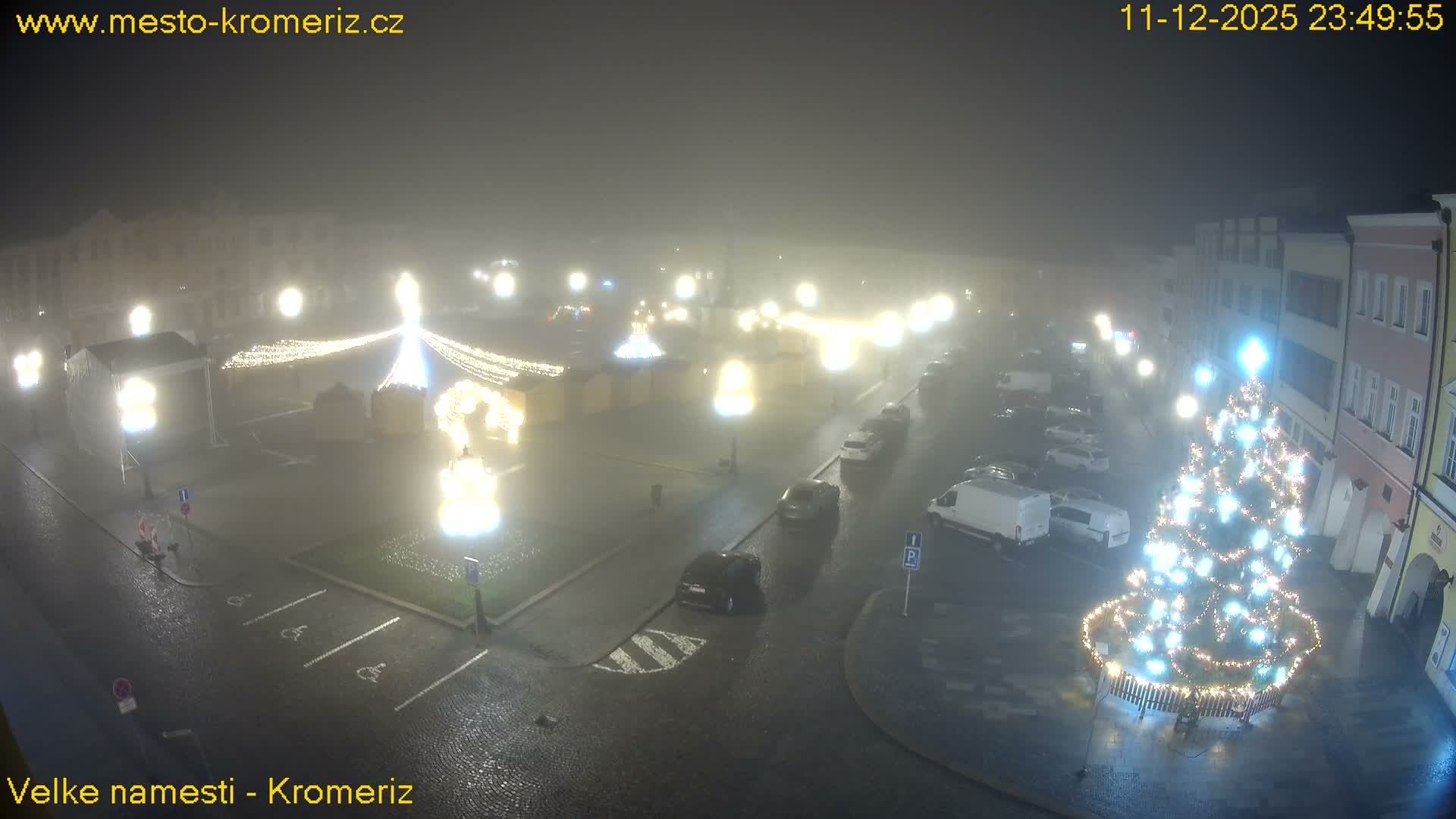 A foggy night view of a festive town square displays multiple brightly lit Christmas trees, market stalls, and parked cars amidst historic buildings.