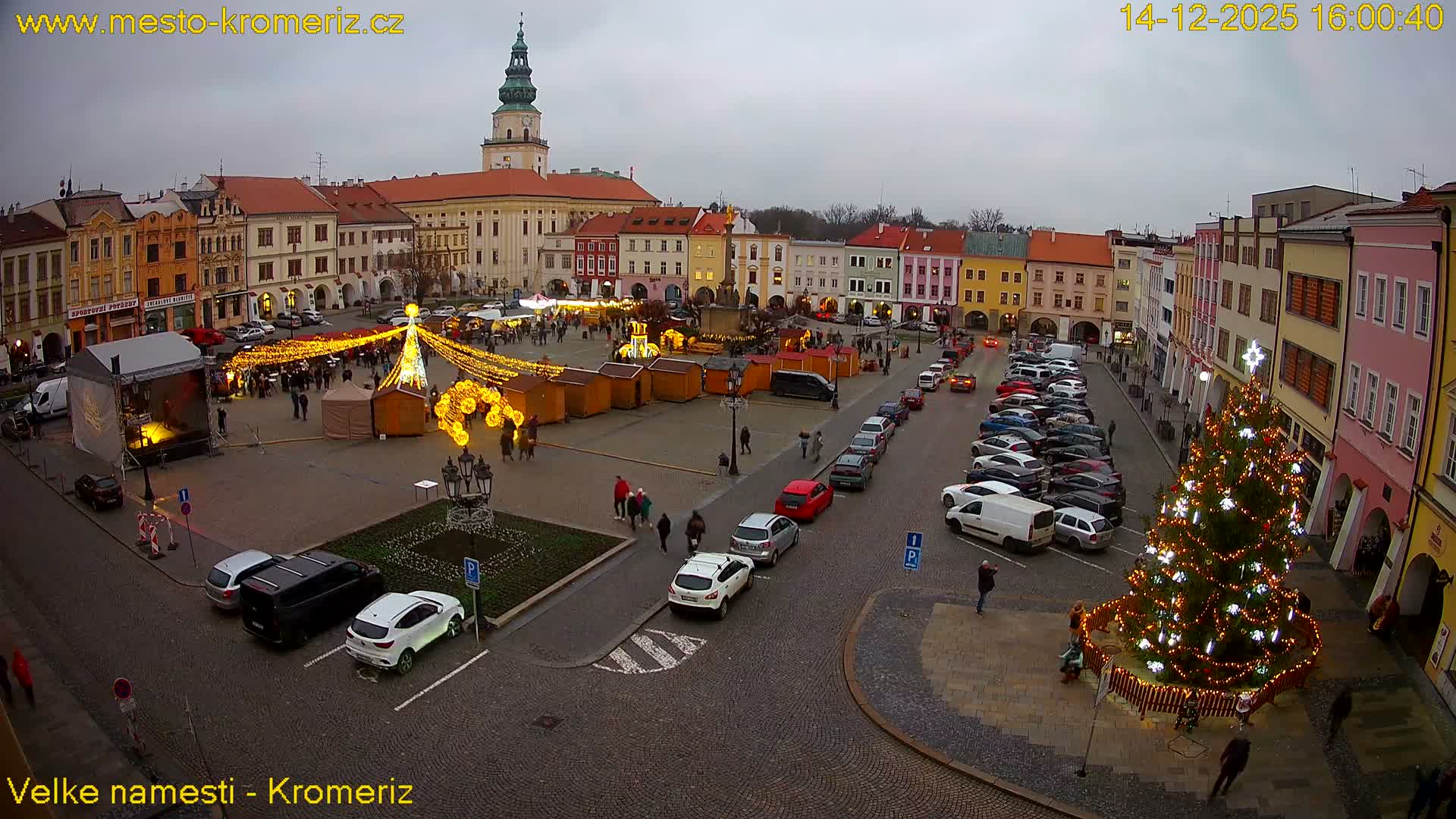 A foggy night view of a festive town square displays multiple brightly lit Christmas trees, market stalls, and parked cars amidst historic buildings.