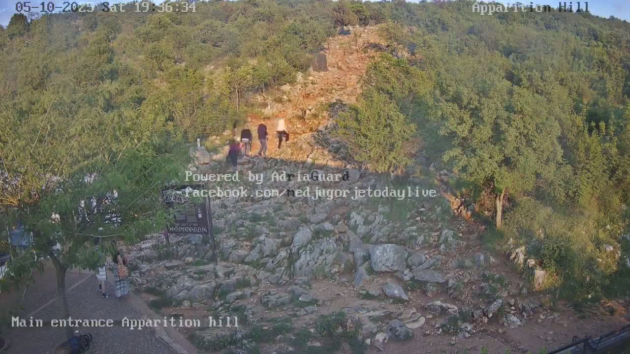A rocky hillside path, partially shaded by trees, is ascended by several people under a clear evening sky.