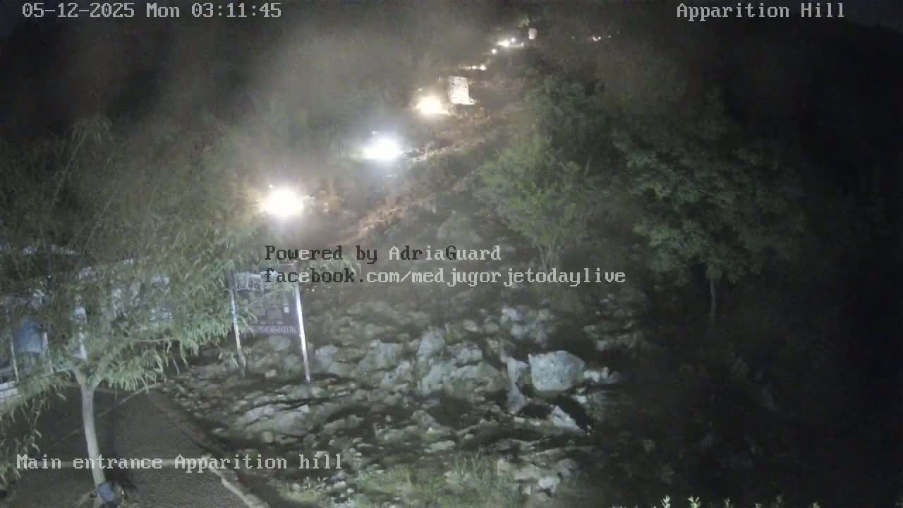 A nighttime, overhead view shows a roadway winding through a rocky, vegetated area with several vehicles visible in the distance.