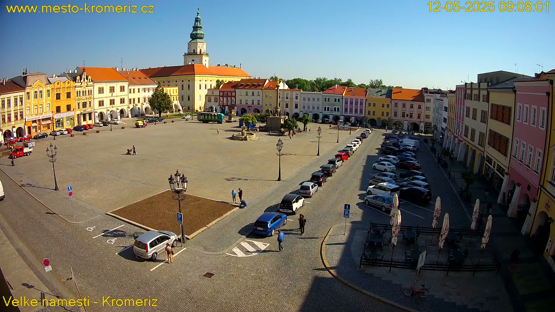 A large town square with colorful buildings surrounding it, a few cars parked along the side, and several people walking around on a sunny day.