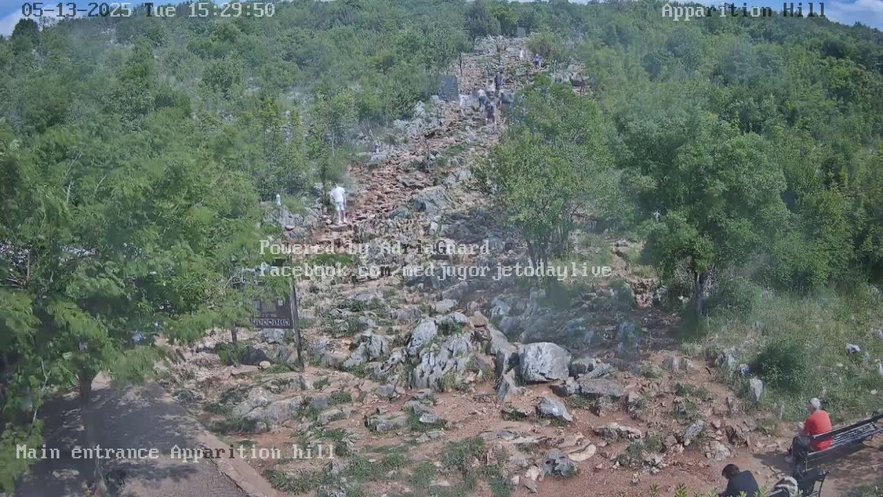 A rocky path winds upward through a green, hilly landscape, with several people walking on it and others sitting near the bottom.