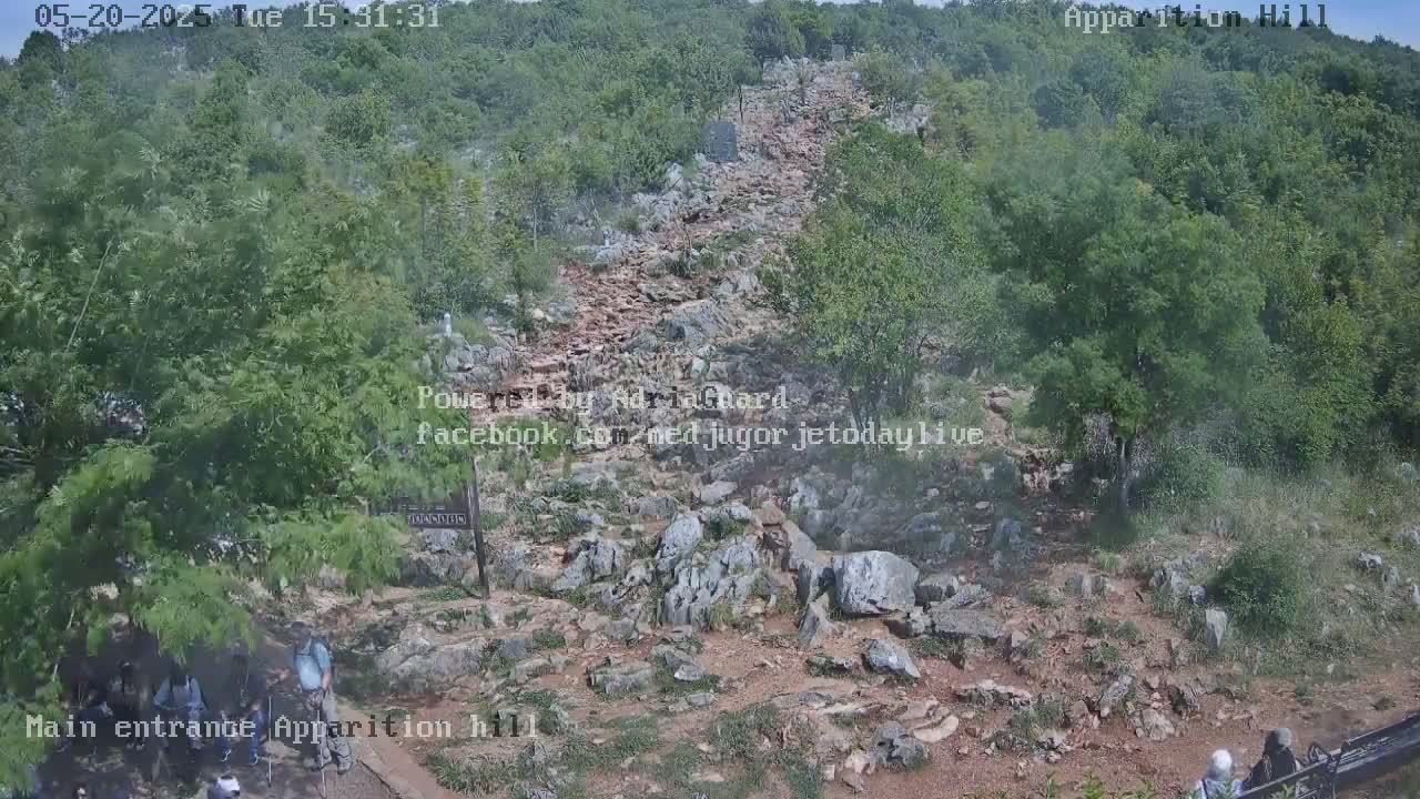 A rocky path winds upward through a hilly, wooded area under a partly cloudy sky.