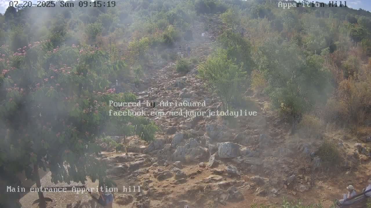 A rocky path winds uphill through hazy, brushy terrain, with a few people visible in the distance.