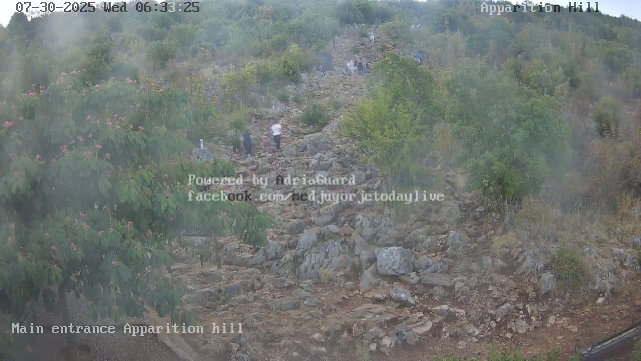 A rocky path winds uphill through hazy, brushy terrain, with a few people visible in the distance.
