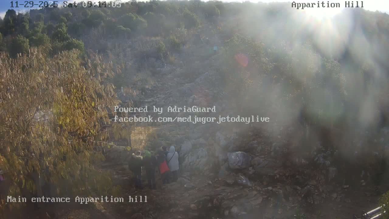 A group of people are gathered on a rocky path winding up a sparsely vegetated hillside under bright, hazy sunshine.