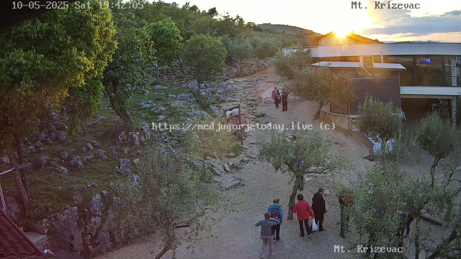 A rocky path winds through a landscape of olive trees and low vegetation towards a modern building at sunset.