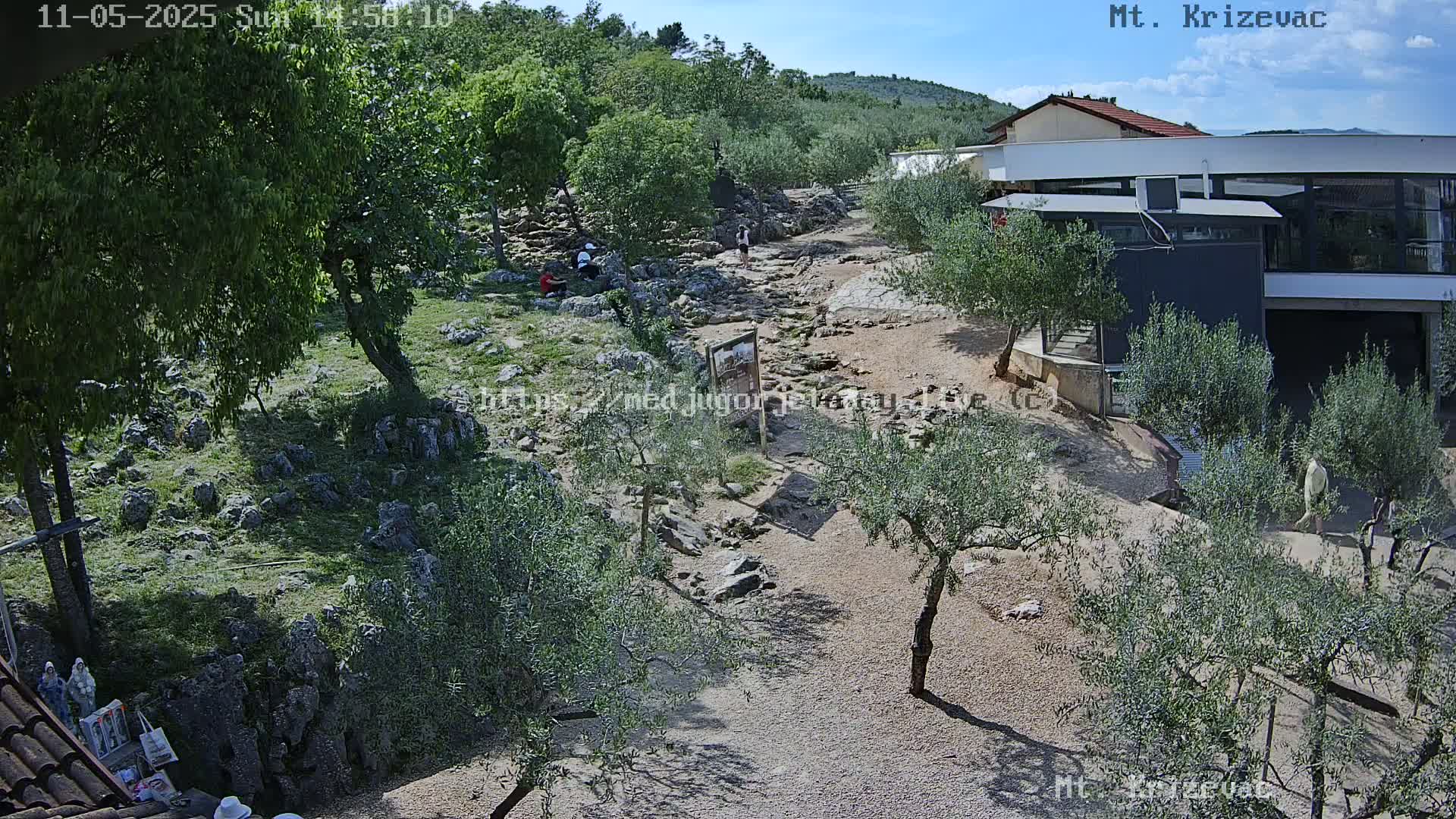 A rocky path winds through a landscape of olive trees and low vegetation under a clear blue sky, with a modern building and a few people visible in the distance.