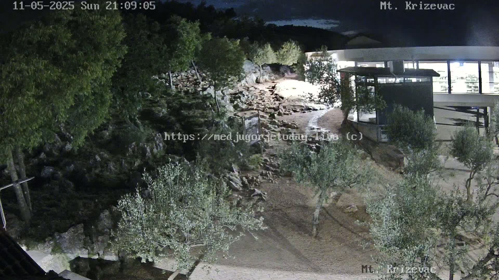 A nighttime, high-angle view shows a rocky, sparsely vegetated area with olive trees and a modern building, illuminated under a dark sky.