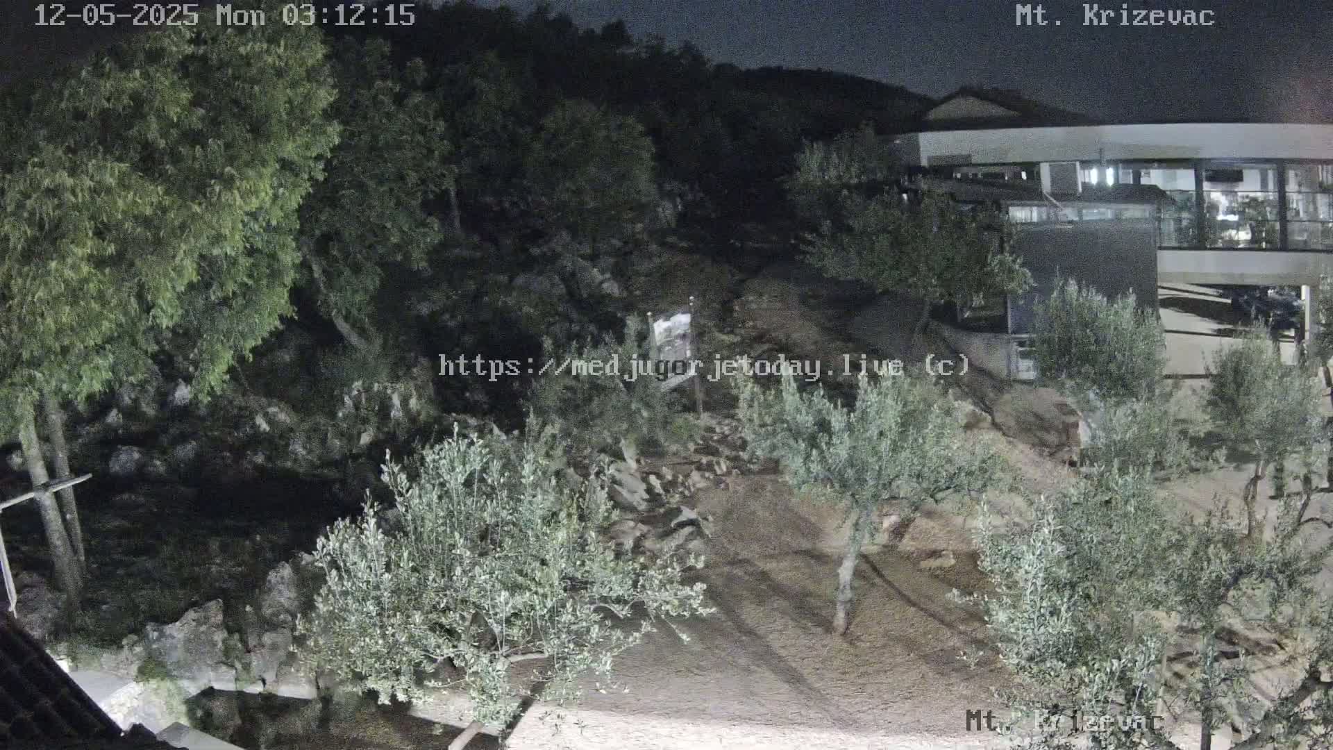 A nighttime view of a rocky hillside with olive trees, a building, and a cross under a dark sky.