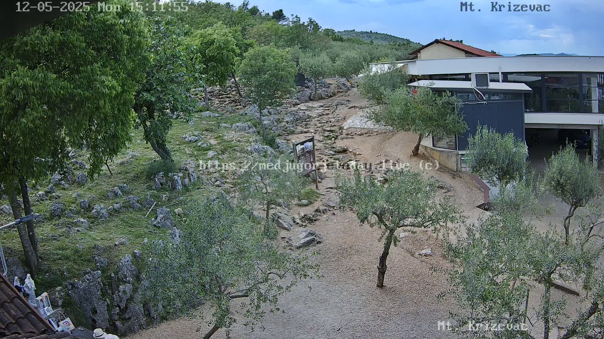 A rocky, grassy area with olive trees and a modern building is visible under a partly cloudy sky.