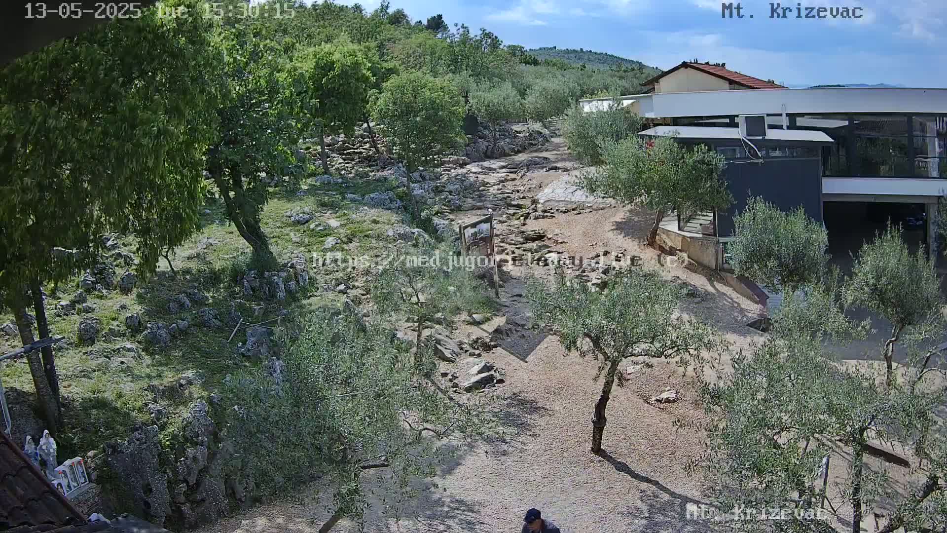 A rocky path winds through a grove of olive trees under a partly cloudy sky toward a modern building, hills and more trees in the background.