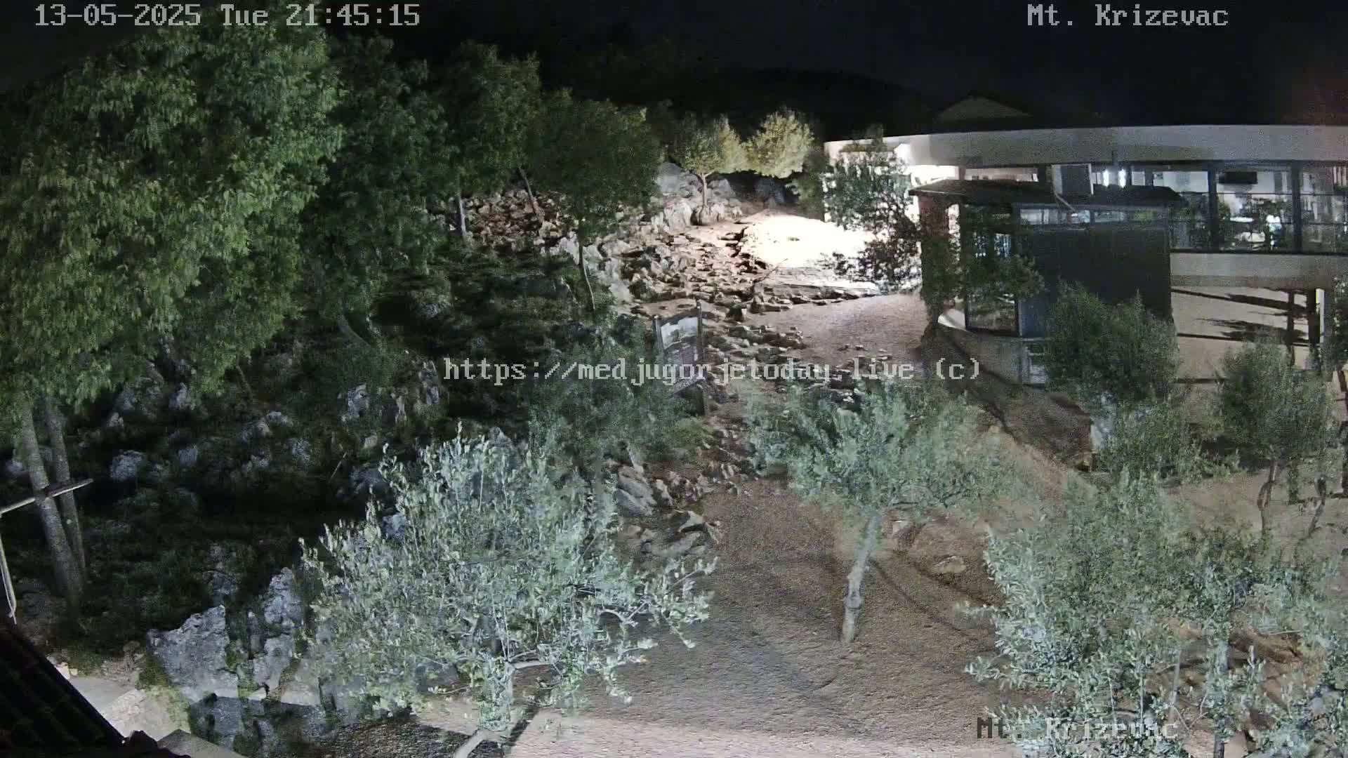 A nighttime, outdoor scene shows a rocky path lit by artificial light, winding through sparse vegetation toward a building with a large, flat roof.