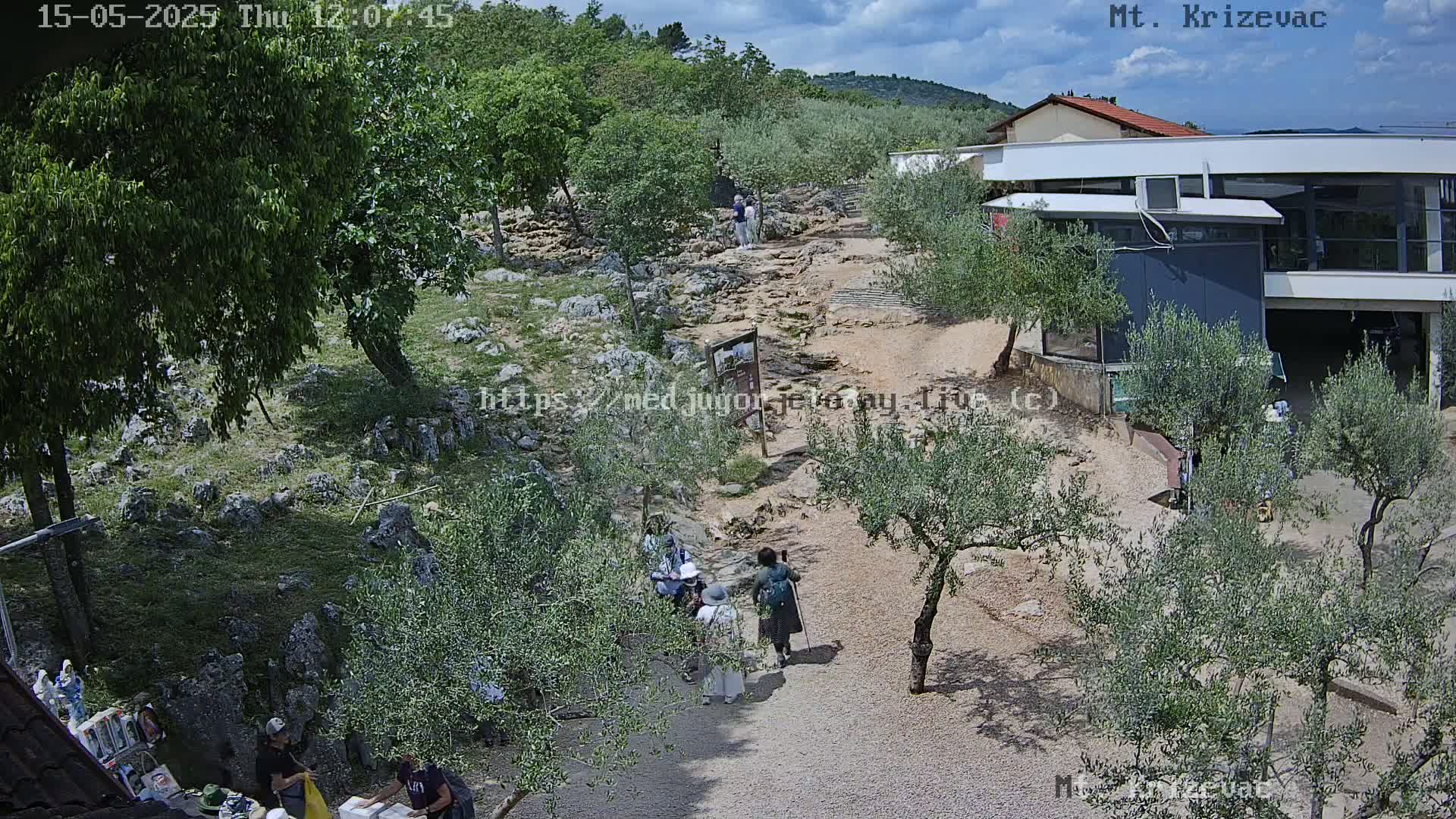 A rocky path winds through a landscape of olive trees and low vegetation under a partly cloudy sky, with a few people walking along the path and a modern building visible in the background.