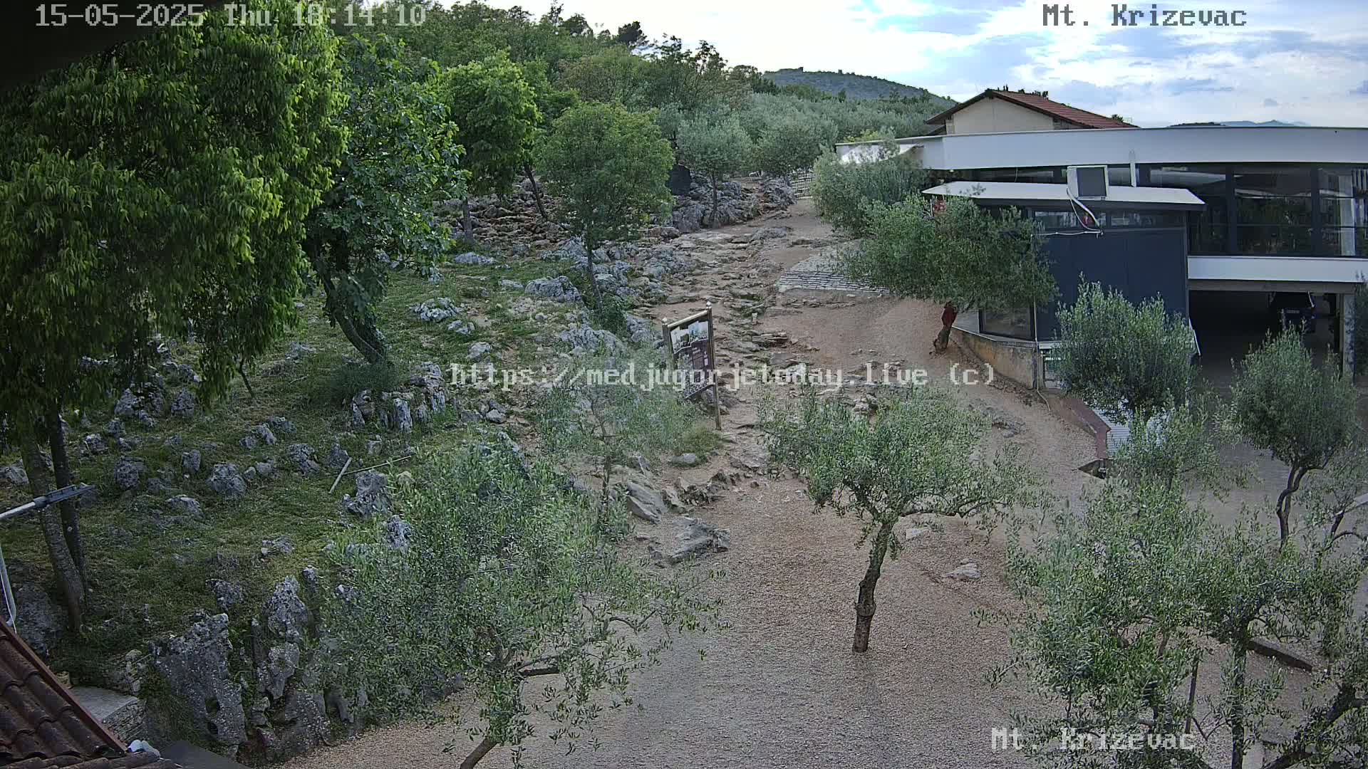 A rocky path winds through a hilly landscape dotted with olive trees under a partly cloudy sky, leading to a building.
