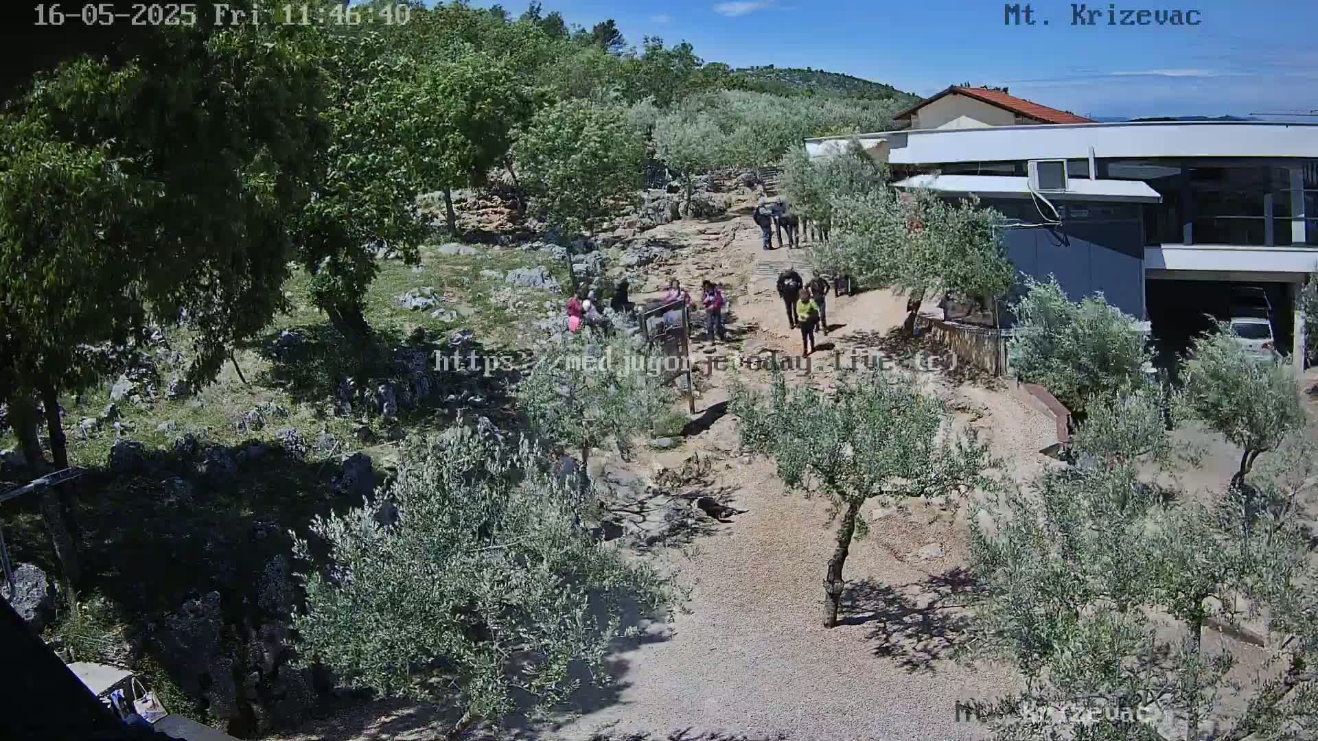 A sunny day reveals a path winding through a rocky, hilly landscape dotted with olive trees and small groups of people walking, leading to a modern building.