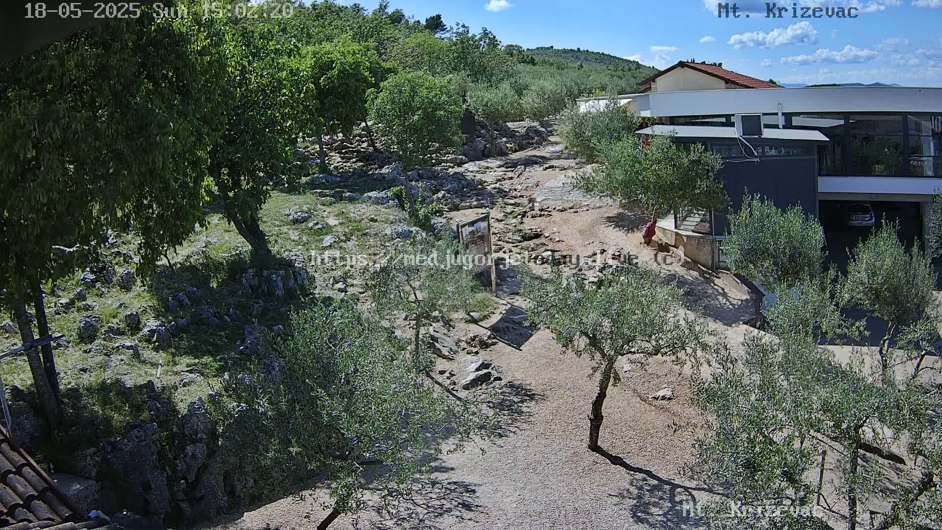 A rocky, gravel path winds through a landscape of olive trees and shrubs under a mostly sunny sky, leading to a modern building.