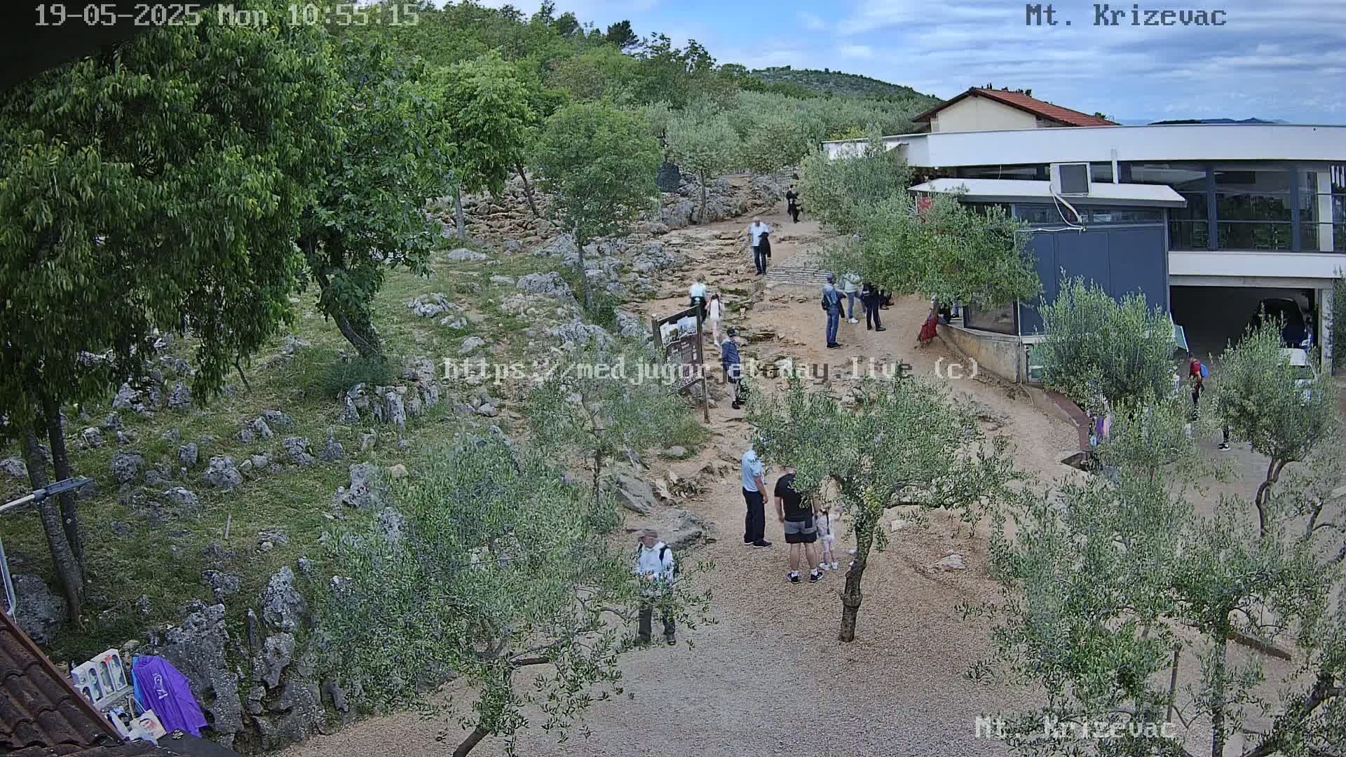 An outdoor scene under partly cloudy skies shows a rocky, gravel path winding through olive groves, with several people walking along it toward a modern building.