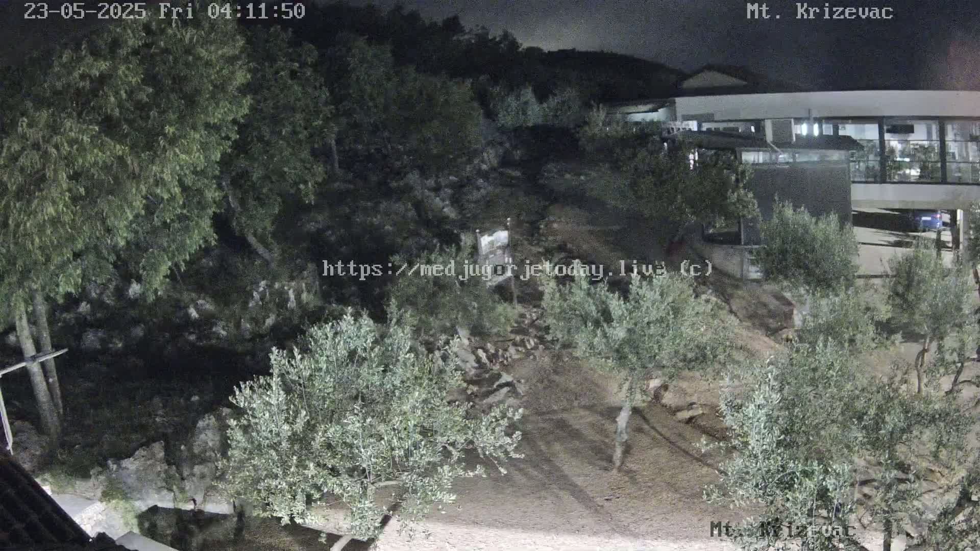 A nighttime, outdoor scene shows a hilltop building with olive trees and rocky terrain under a dark sky.