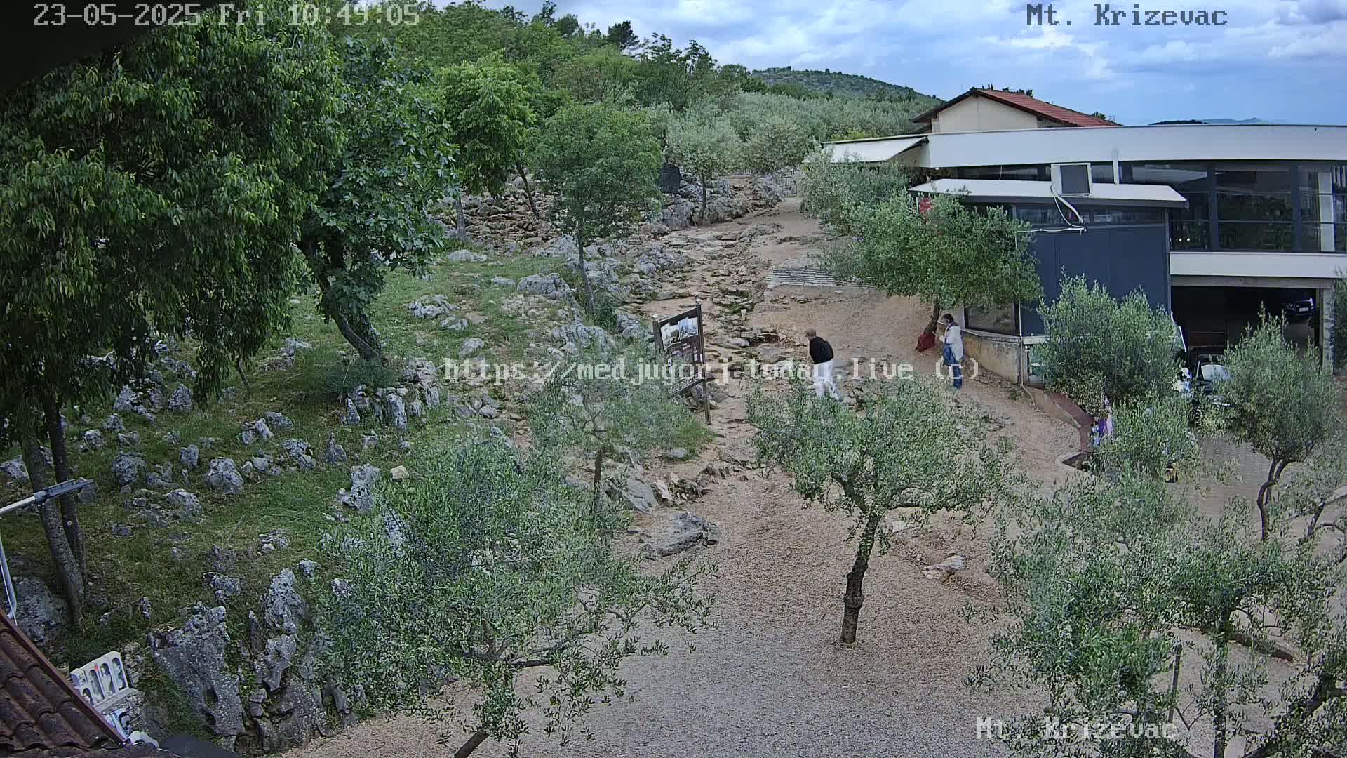 A rocky, olive tree-lined path winds toward a modern building under a partly cloudy sky.