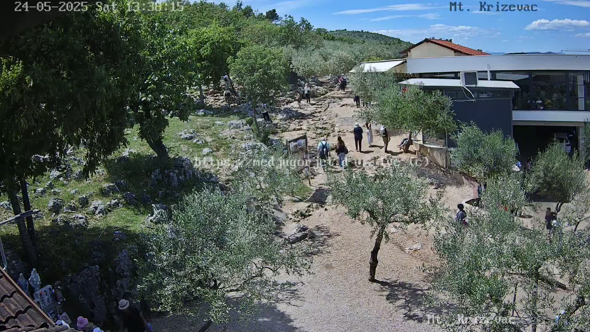 A rocky, sunlit path winds through a landscape of olive trees and low vegetation toward a modern building, with numerous people walking along the path under a partly cloudy sky.