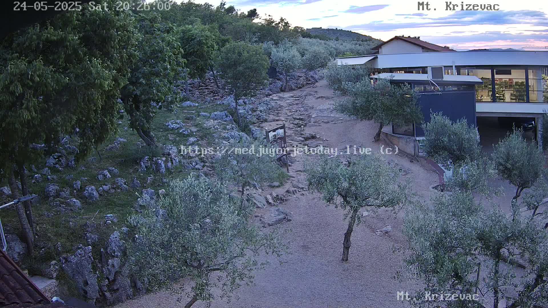 A rocky path winds through a landscape of olive trees and low vegetation toward a modern building under a twilight sky.