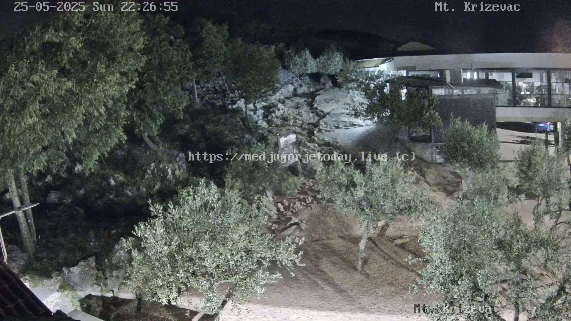 A nighttime, high-angle view shows a rocky hillside with olive trees, a modern building, and a parking lot.