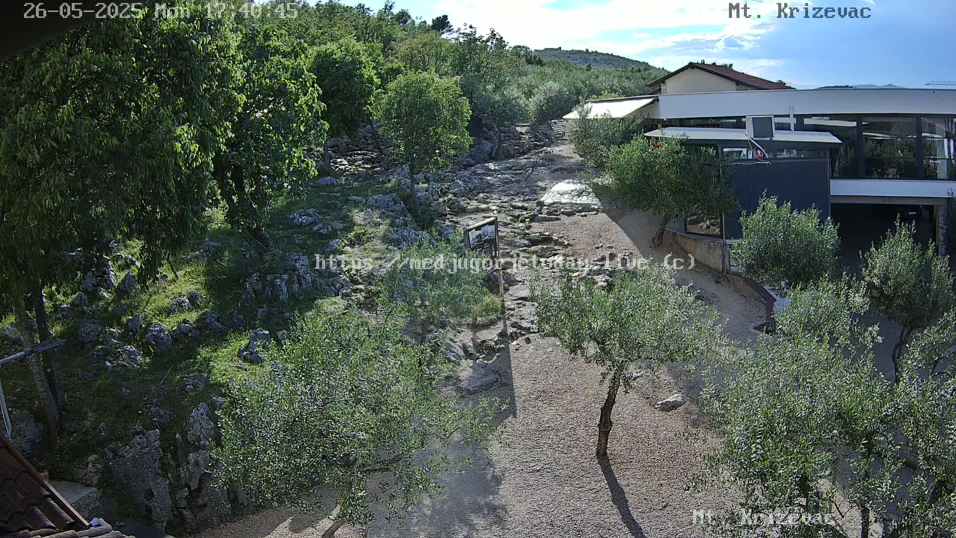 A rocky, partially paved path winds through a landscape of olive trees and low-lying vegetation under a mostly sunny sky.
