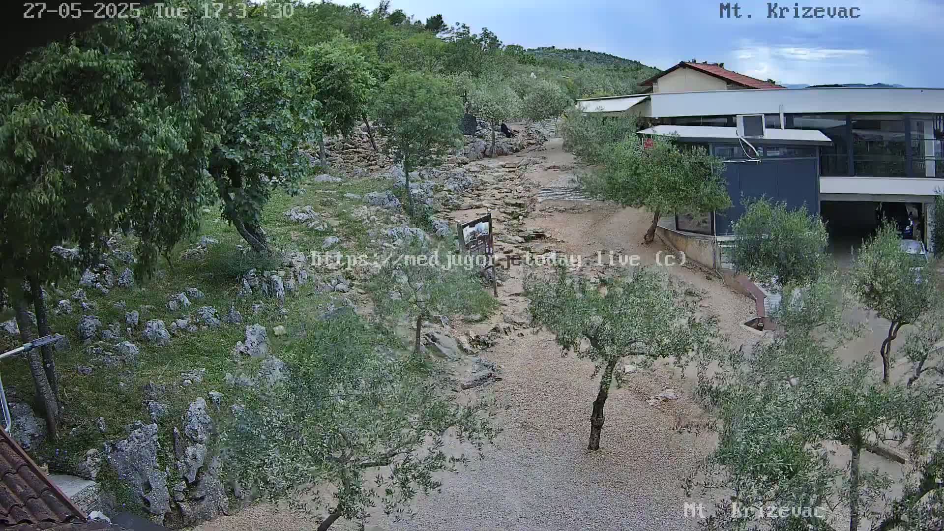 An overhead view of a rocky, dry creek bed meandering through a landscape of olive trees and stone structures under a partly cloudy sky.