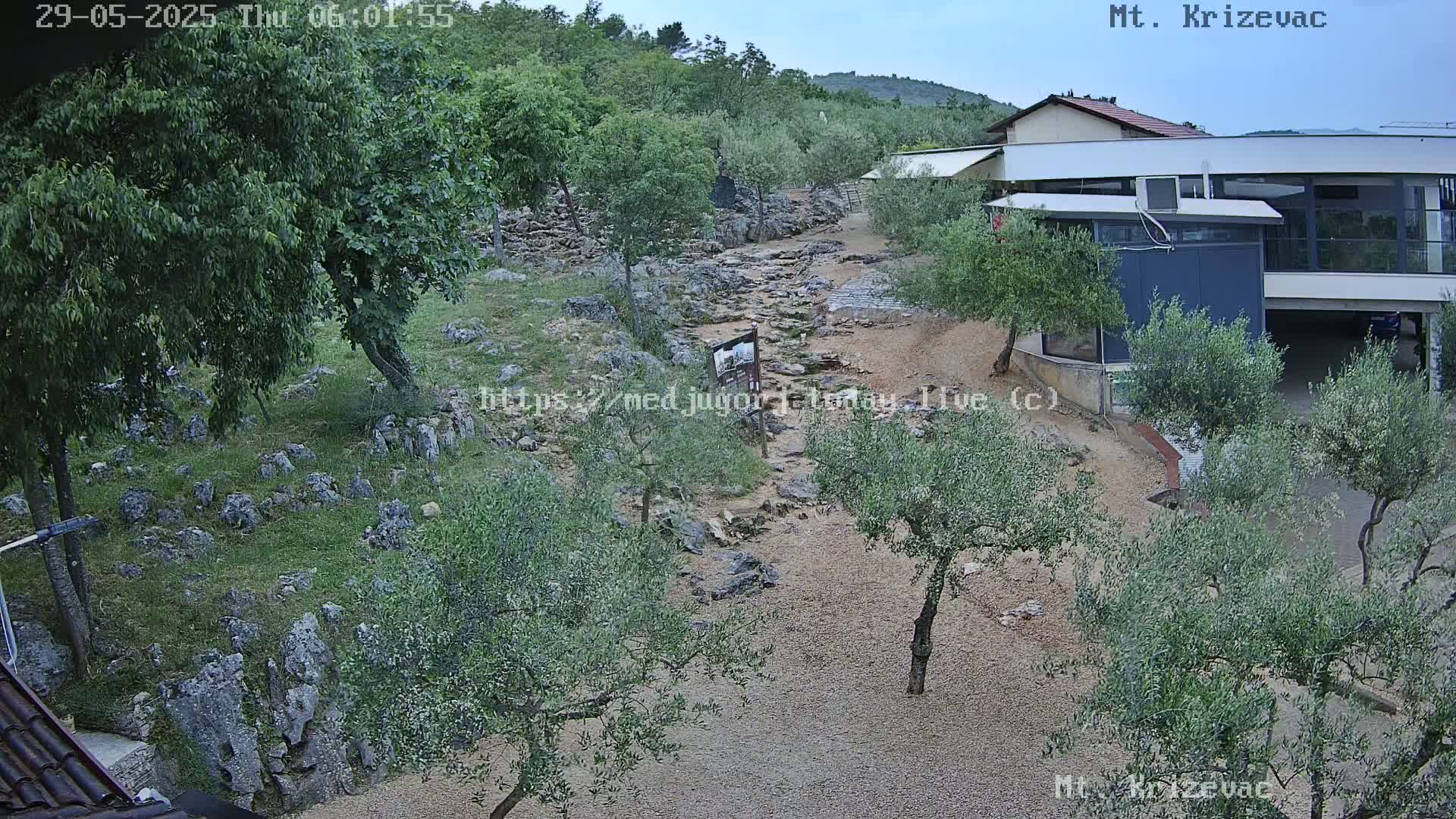 A rocky, dry creek bed winds through a landscape of olive trees and low vegetation toward a modern building under an overcast sky.