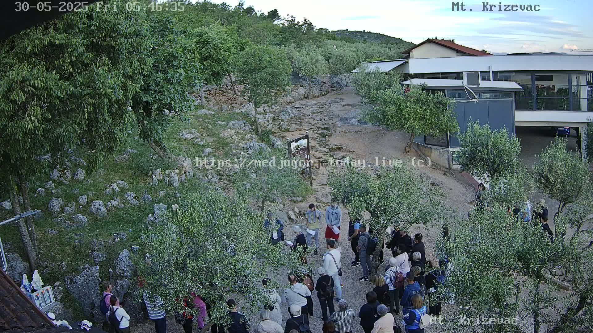 A large group of people gathers outdoors in a rocky area with olive trees and a modern building under a partly cloudy sky.