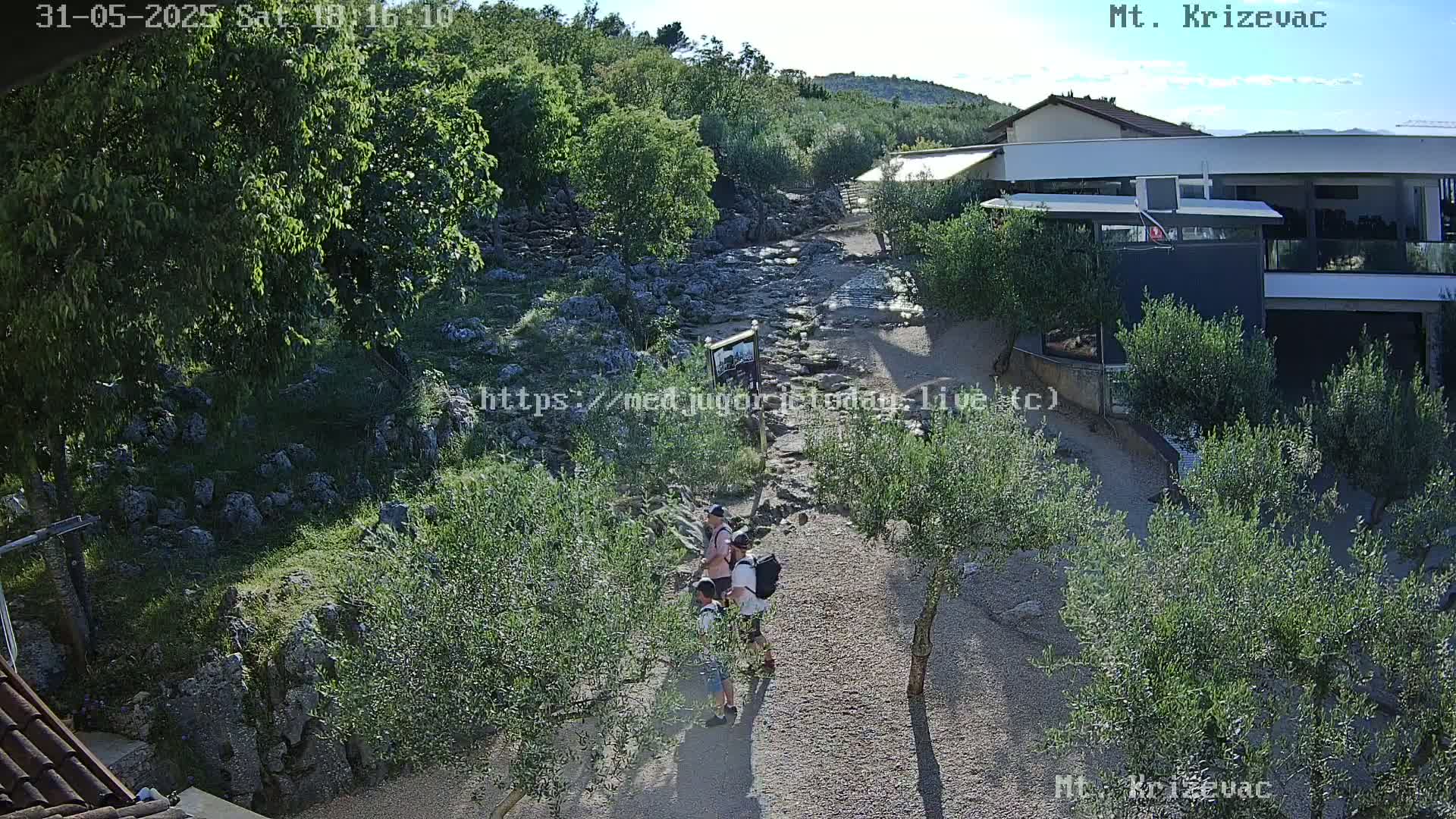 A sunny outdoor scene shows a dry creek bed with a few people walking on a path nearby, surrounded by olive trees and a building in the background.