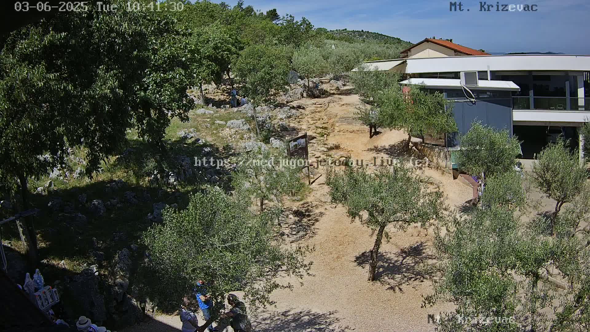 A sunny outdoor scene shows a rocky path winding through olive trees towards a modern building, with several people walking along the path.