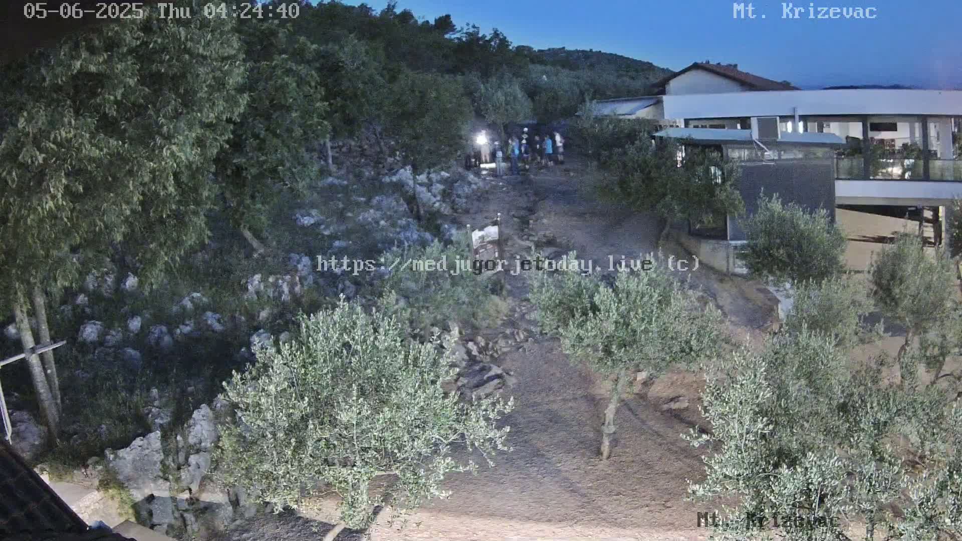 A nighttime outdoor scene shows a group of people standing near a rocky hillside with olive trees and a building in the background under a twilight sky.