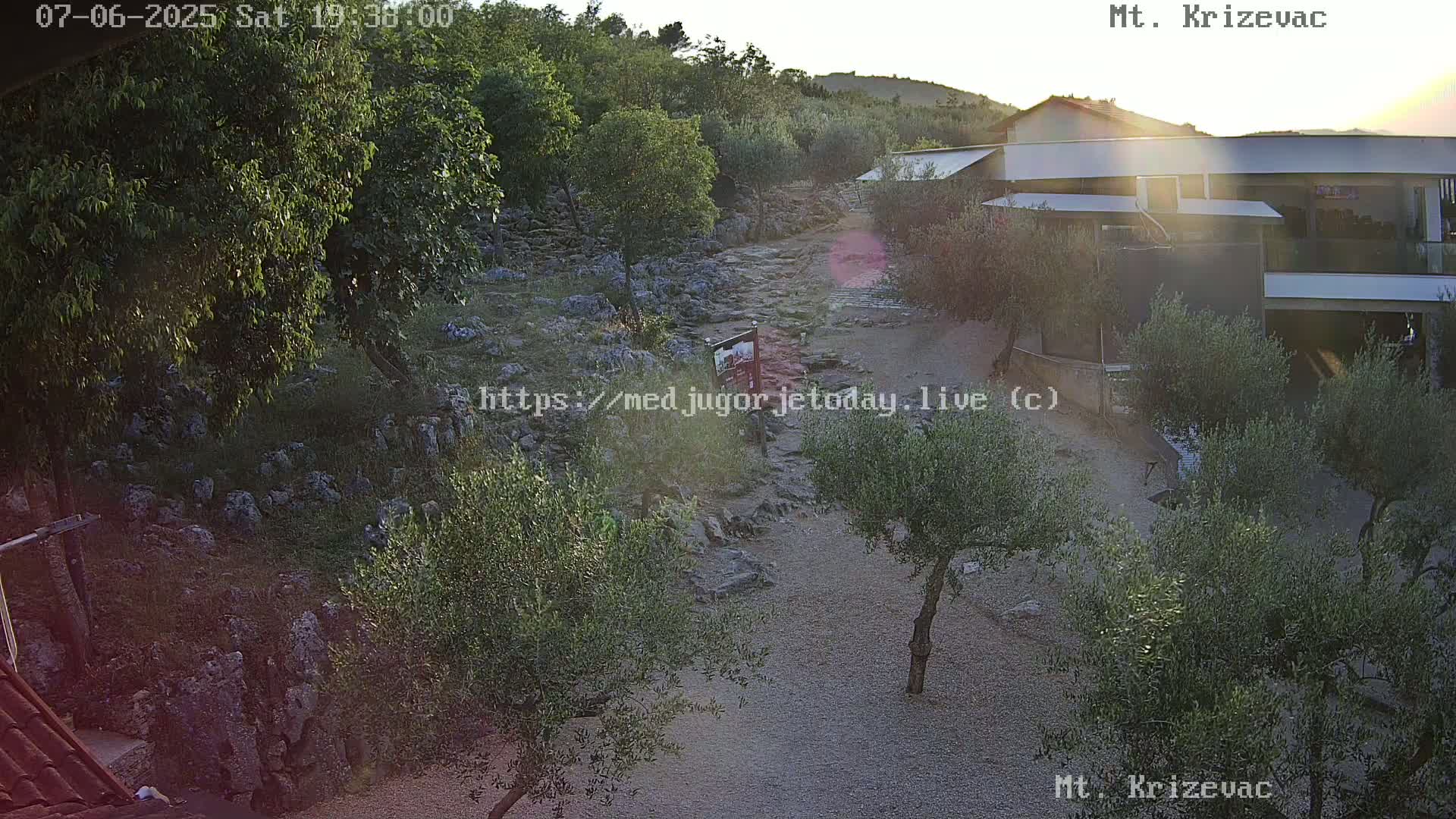 An elevated view shows a rocky path winding through a sun-drenched landscape dotted with olive trees and a modern building, under a clear sky at sunset.