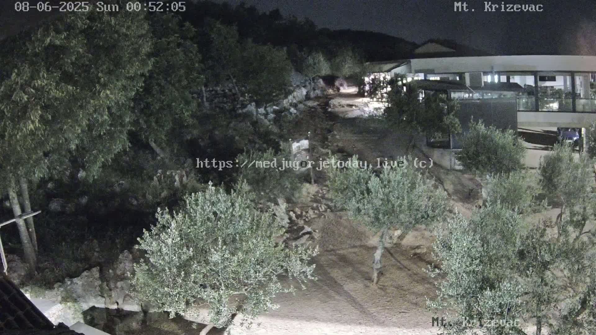 A nighttime, outdoor view shows a rocky area with olive trees, a building, and a cross.