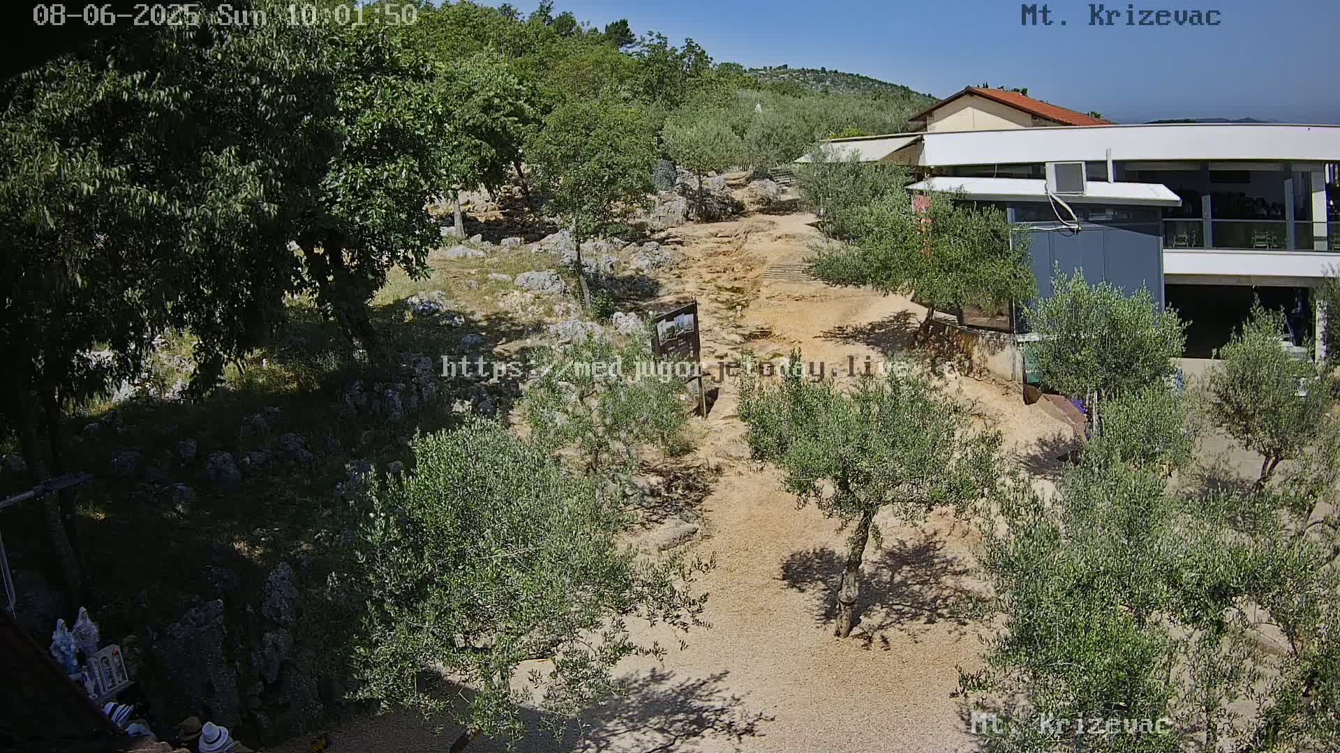 A sunny outdoor scene shows a light-brown dirt path winding through olive trees toward a modern building nestled among hills.