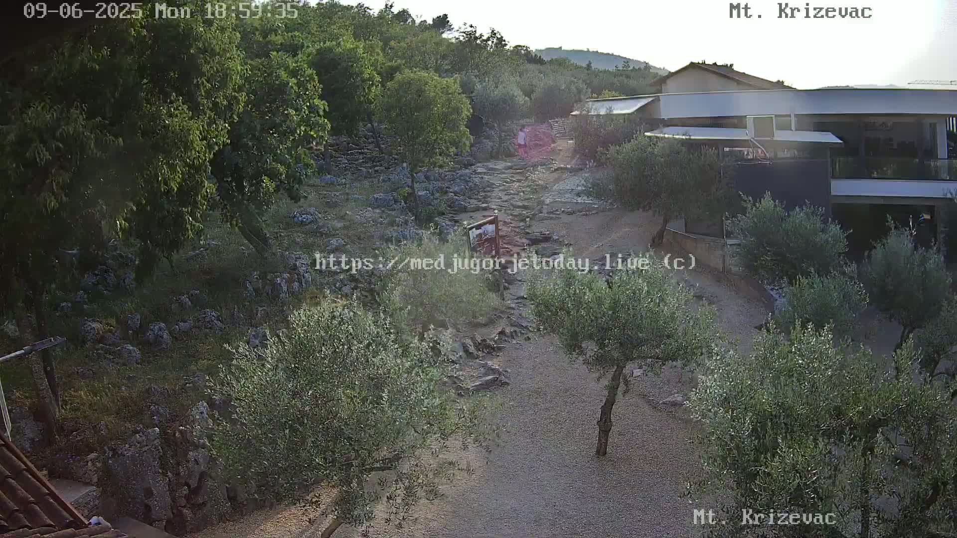 A rocky path winds through a landscape of olive trees and low buildings under a clear, bright sky.
